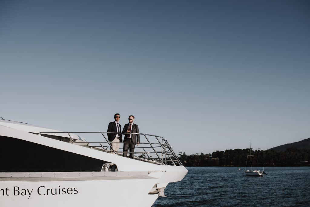 Two men dress in suits standing on the bow of a white catamaran
